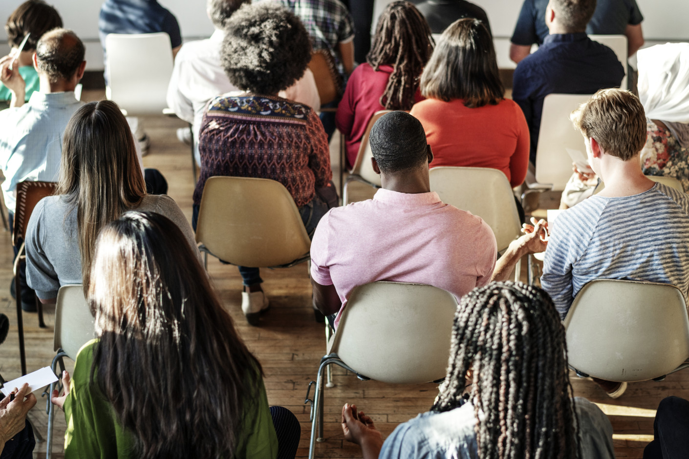 A group of people sit in rows of chairs in a bright room, seen from behind as they face a speaker.