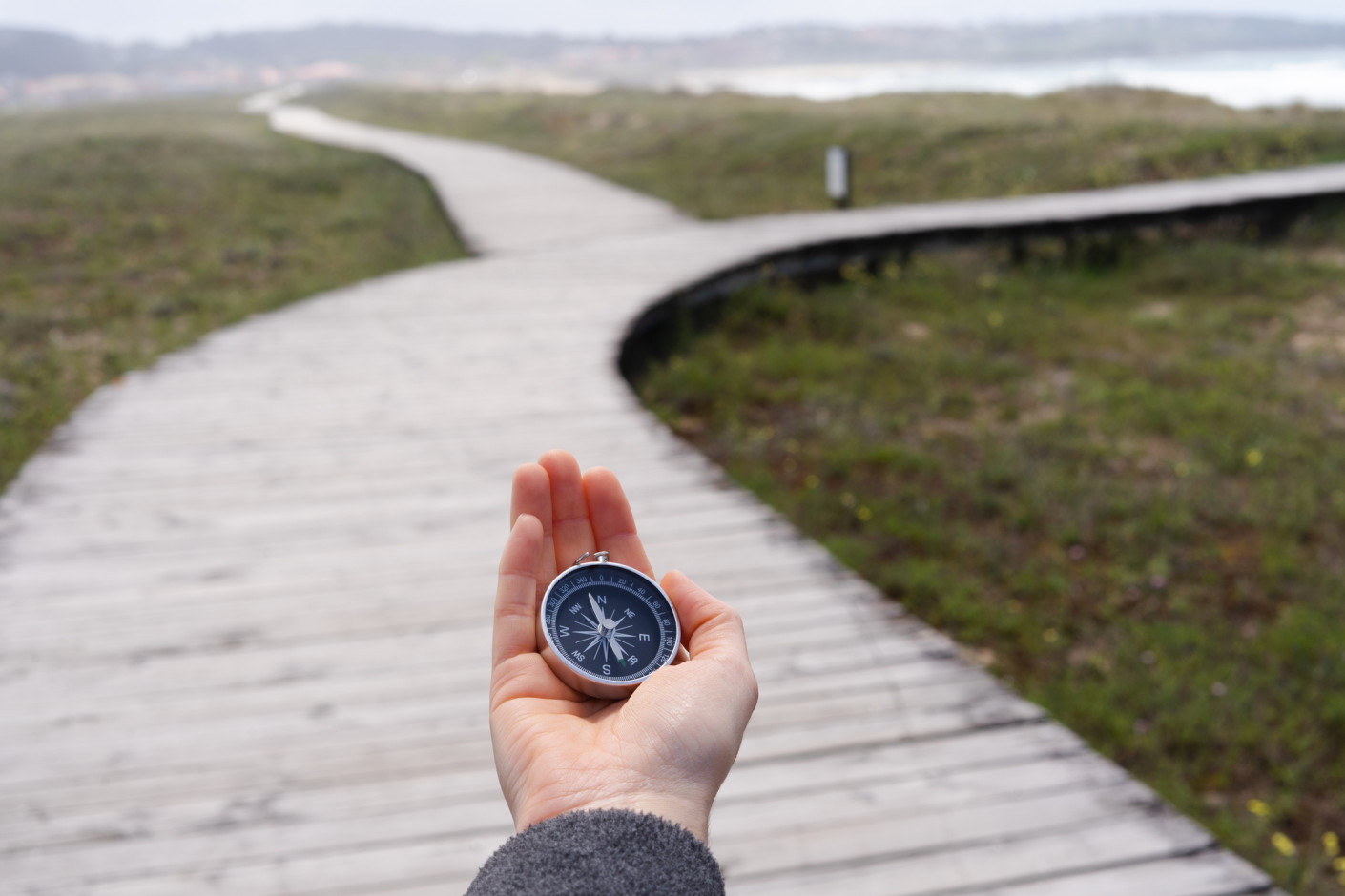 A hand holds a compass in the foreground of a wooden boardwalk curving through green marsh toward water.