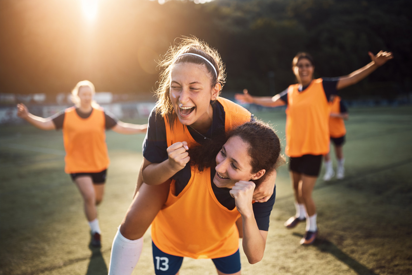 Women in orange jerseys celebrate on a grass field, two in front embracing while others run behind them.