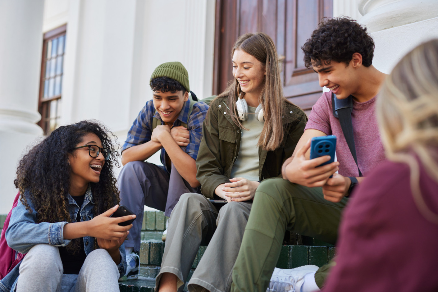Four young people sit on outdoor steps, smiling and looking at phones together.