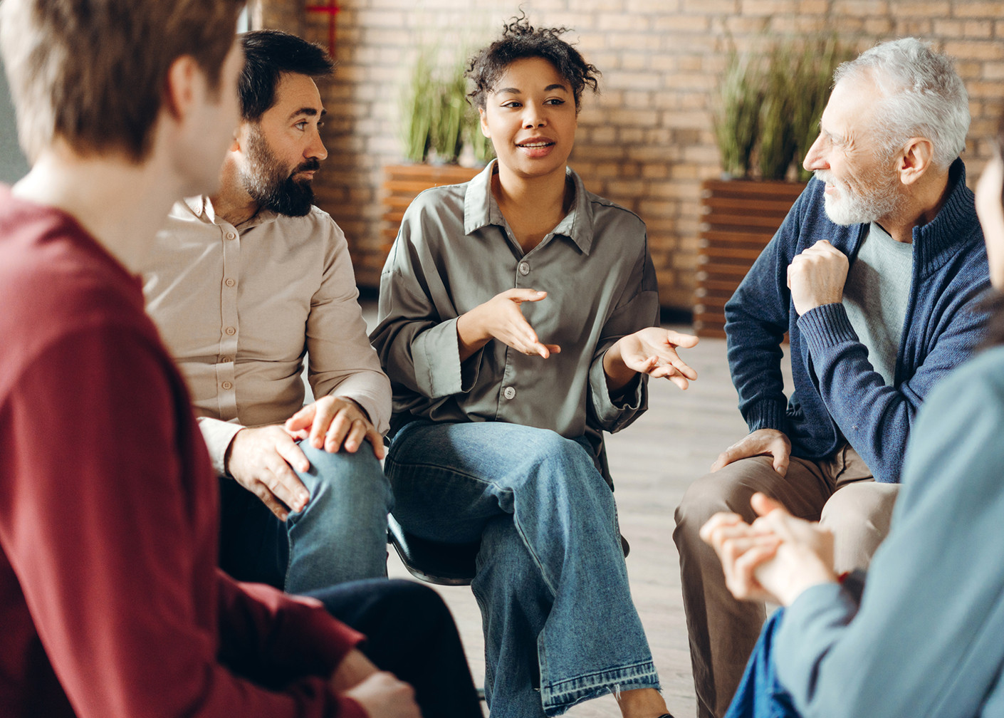 People sit in a circle of chairs indoors, talking, with one person gesturing.