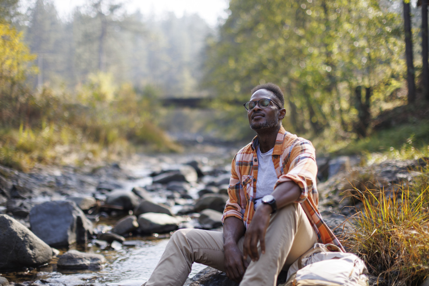A man in a plaid shirt and glasses sits on a rock beside a shallow stream in a forest.